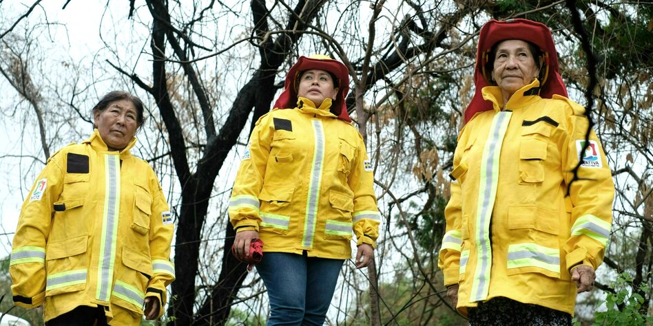 Drei Frauen in gelber Feuerwehruniform stehen vor abgebrannten Bäumen