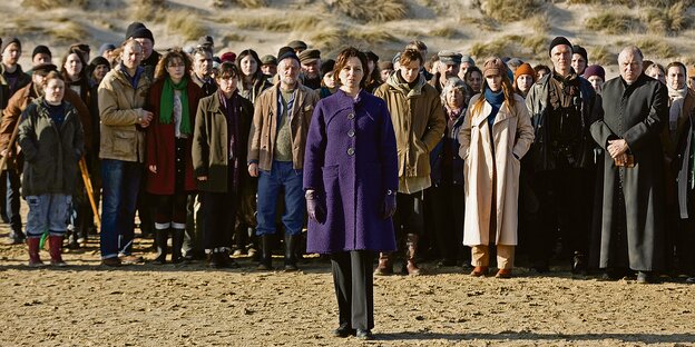 Eine Frau steht im Sand vor einer Ansammlung von Menschen. Alle schauen in die gleiche Richtung. Im Hintergrund ist eine Düne.