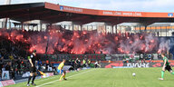 Fans auf einer Stadiontribüne brennen rote Pyros ab