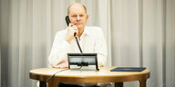 Olaf Scholz sitzt an einem kleinen ovalen Tisch vor einem hellen Vorhang, er hält den Telefonhörer ans Ohr und schaut ernst in die Kamera